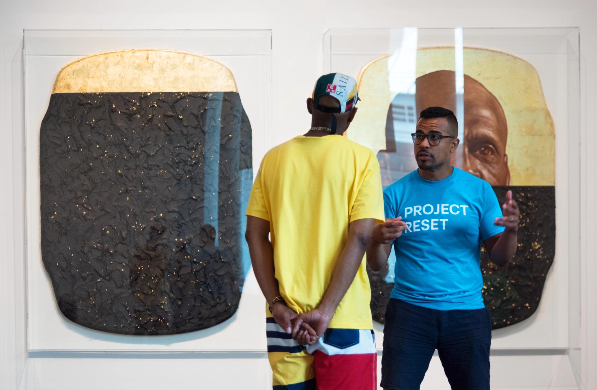 Person in a blue "Project Reset" t-shirt speaks to another person facing a large, gilded artwork