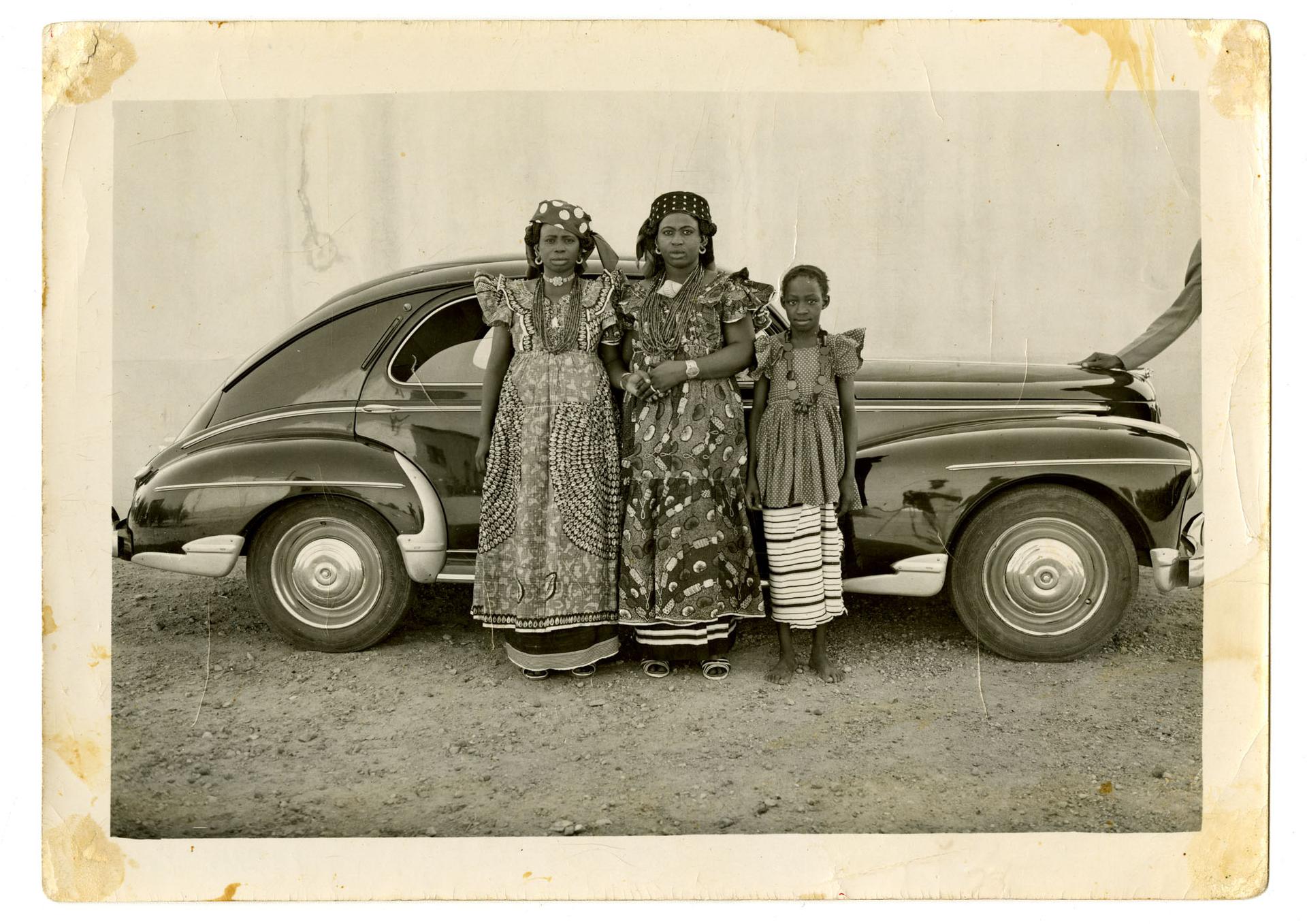 Photograph of three people in patterned clothing standing beside a vintage car