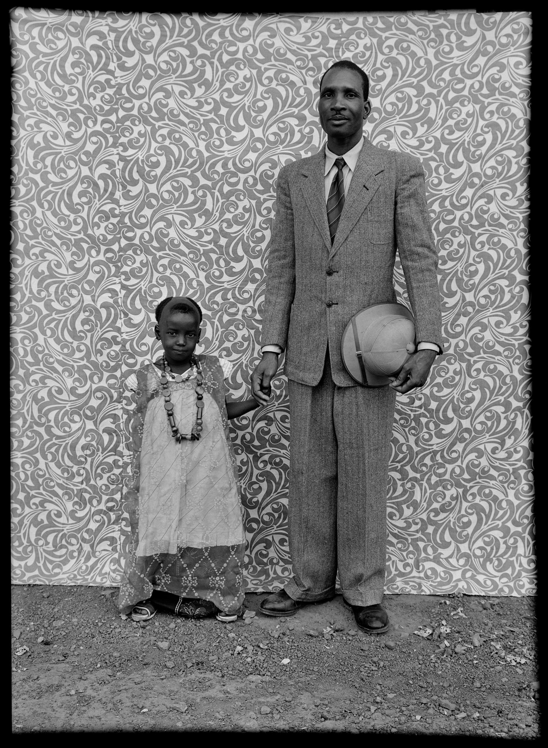 Black-and-white photograph of a man and young child holding hands in front of a patterned backdrop