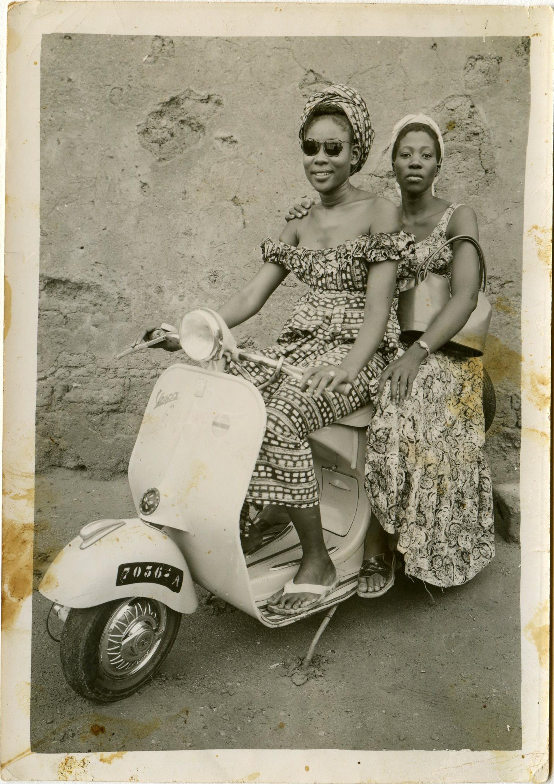 Photograph of two women on a Vespa, both in patterned dresses