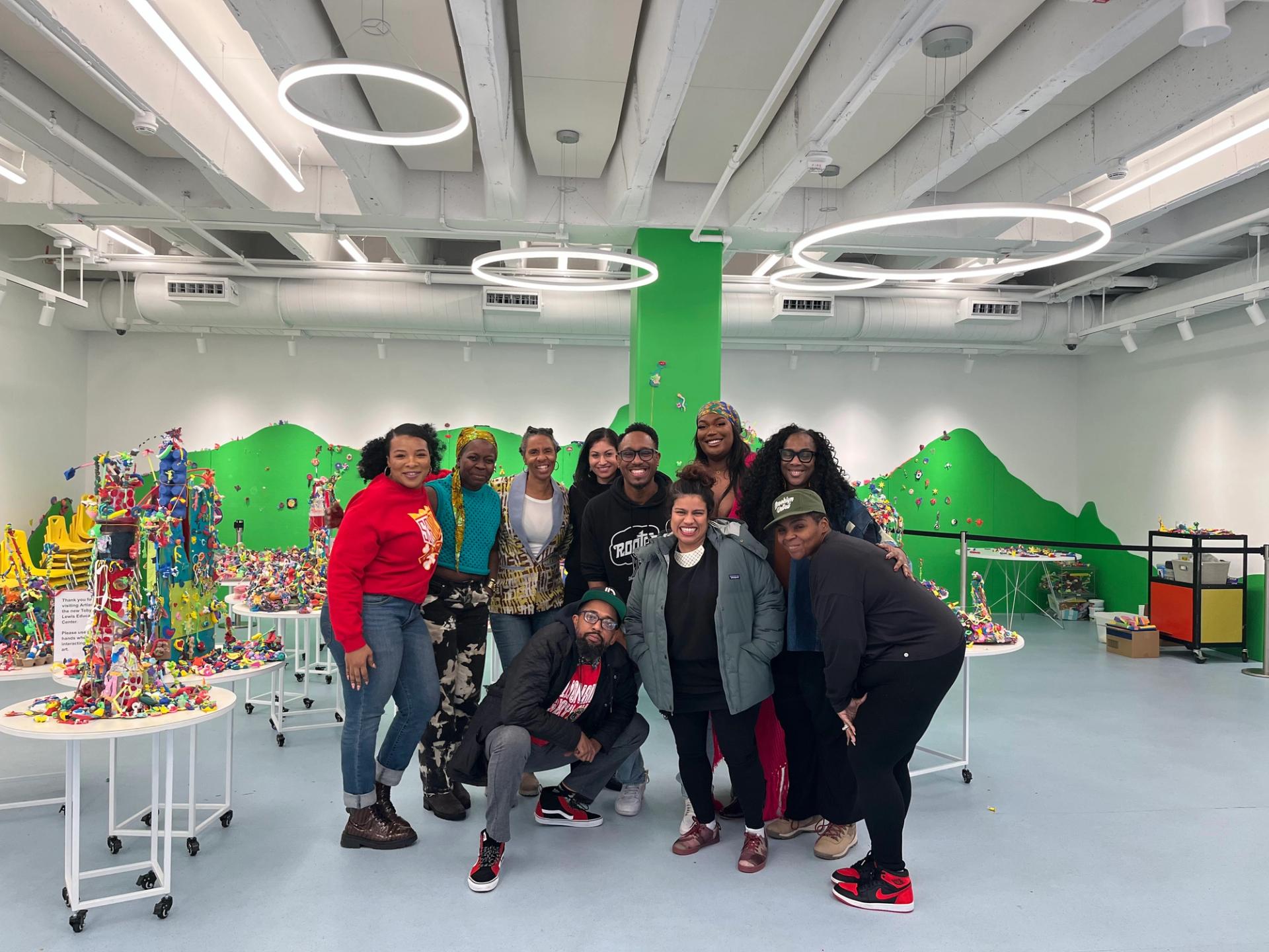 A group of ten adults pose together in a large bright room with green walls, and tables of playdough sculptures