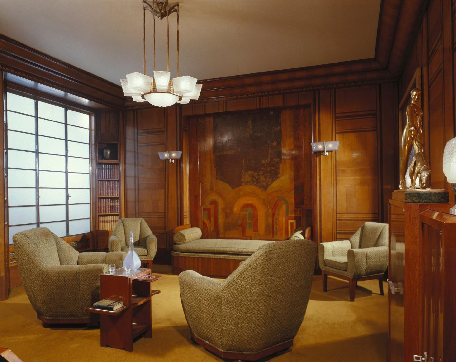 An Art Deco lounge with wood-paneled walls, plush armchairs, a daybed, and decorative sculptures