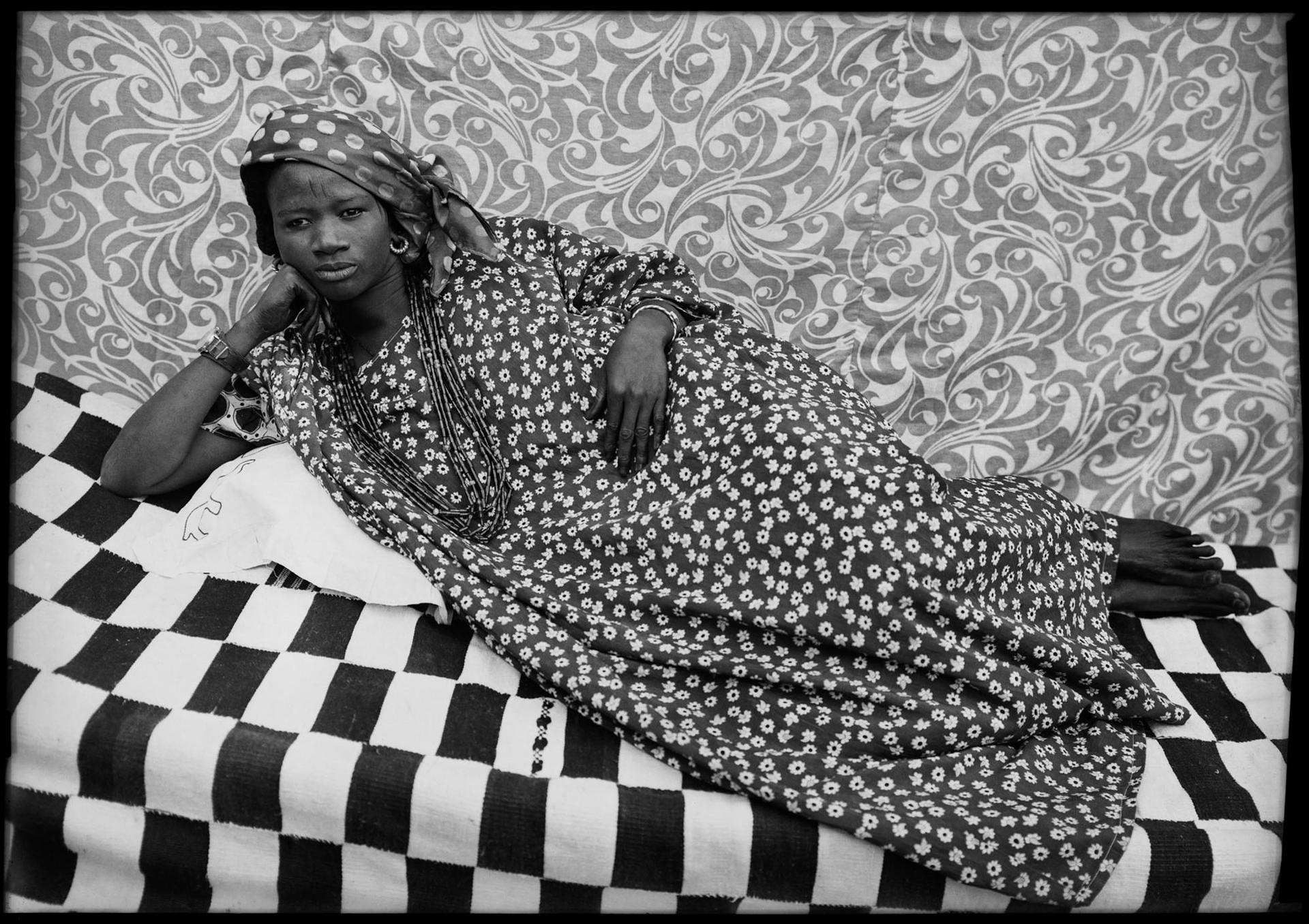 Black-and-white photograph of a woman in a patterned dress reclining on a checkered cloth