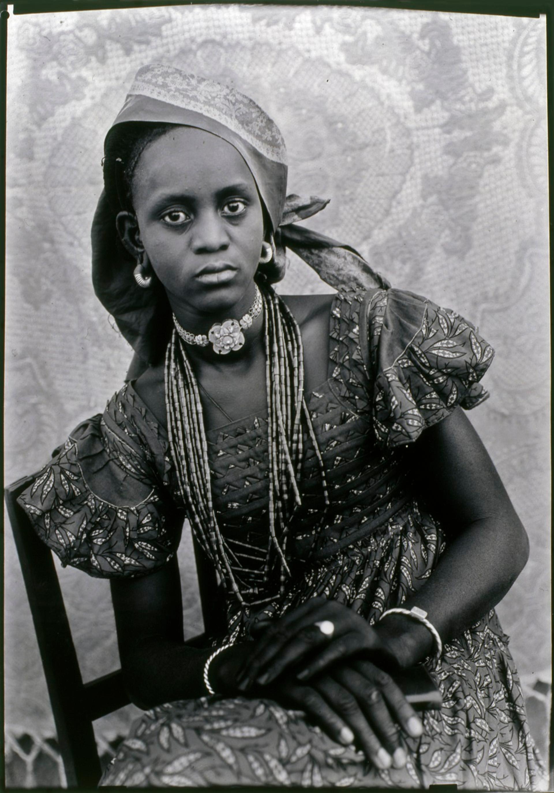 Black-and-white photograph of a woman wearing a patterned dress, flower choker, and headscarf